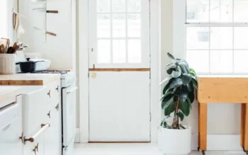 A imagem mostra uma cozinha minimalista toda em branco. Os móveis de madeira presentes são armários (em branco) e uma mesa retrátil (em tom de madeira). Há também um fogão, utensílios como faca e panela, além de uma planta em um vaso branco.