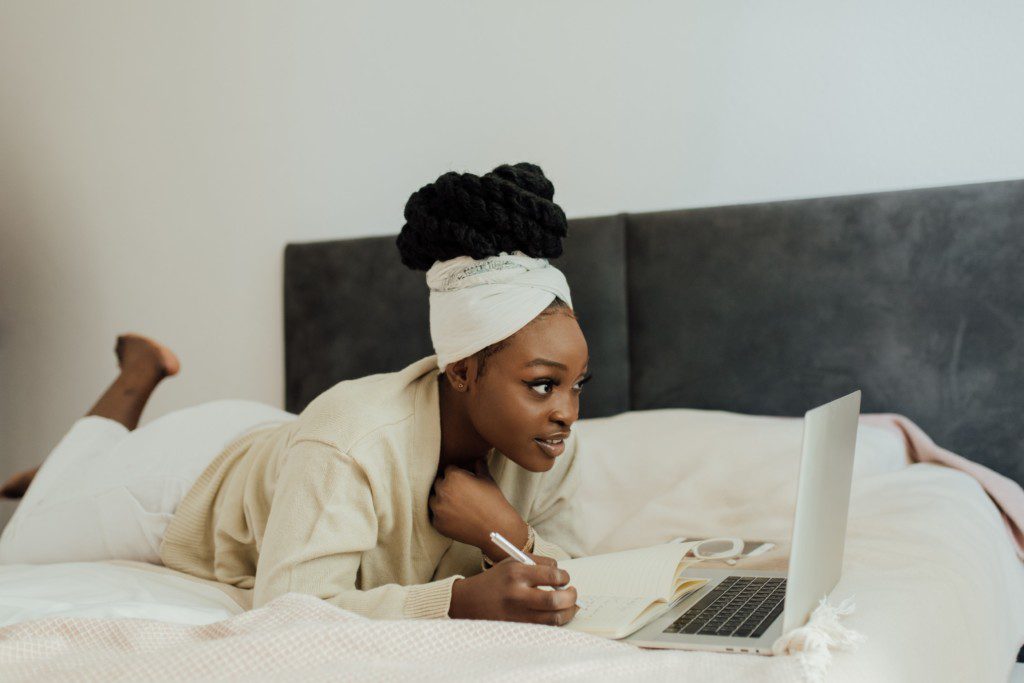 Foto que ilustra matéria sobre receber amigos em casa mostra uma mulher deitada na cama olhando para o computador e fazendo uma lista anotando em um caderno