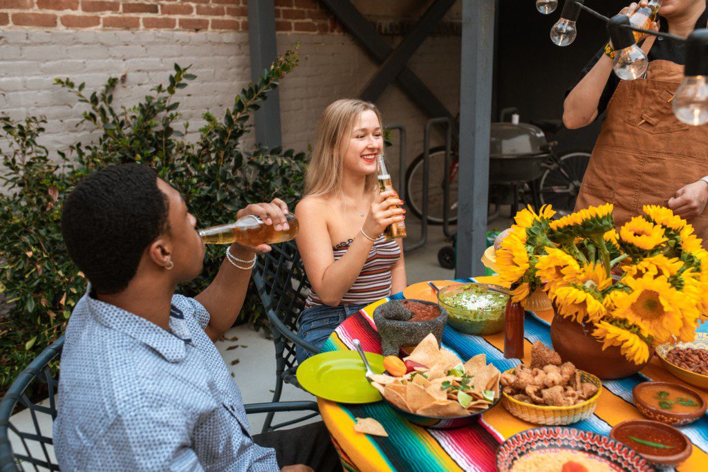 Foto que ilustra matéria sobre como receber amigos em casa mostra duas pessoas sentadas à mesa, com petiscos e bebidas, e um arranjo de flores no centro