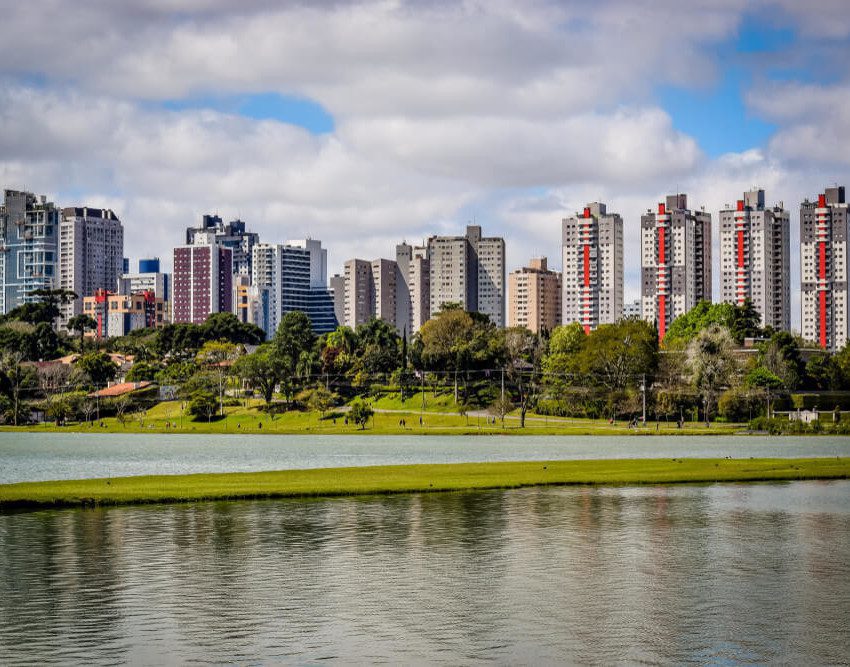 Foto que ilustra matéria sobre qualidade de vida em Curitiba mostra uma panorâmica do Parque Barigui, com um espelho d’água em primeiro plano, árvores no meio da imagem e altos prédios ao fundo