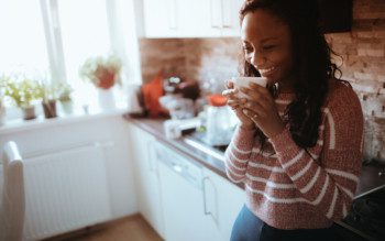 Mulher segurando uma xícara e sorrindo encostada em balcão do cantinho de café de sua casa