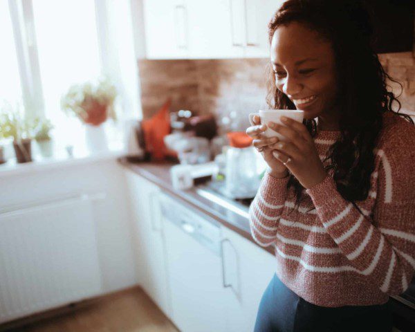 Mulher segurando uma xícara e sorrindo encostada em balcão do cantinho de café de sua casa