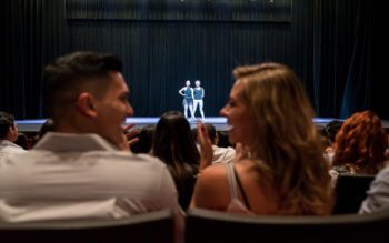 Casal observa fim de espetáculo em um teatro com casa cheia em São Paulo. Ao redor, todos aplaudem e o casal ri, dando a ideia de que gostou do show