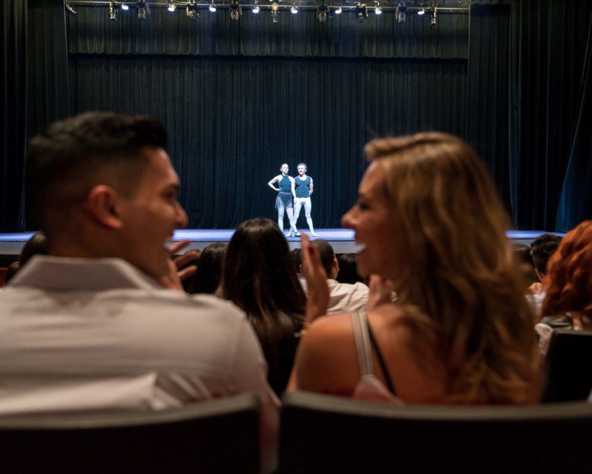Casal observa fim de espetáculo em um teatro com casa cheia em São Paulo. Ao redor, todos aplaudem e o casal ri, dando a ideia de que gostou do show
