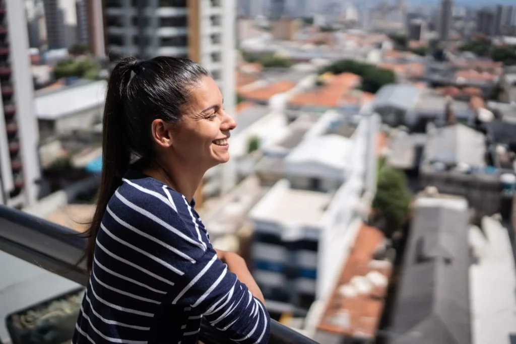 Imagem de uma mulher sorrindo ao contemplar a cidade de São Paulo vista do alto de um apartamento para ilustrar matéria sobre a segunda via da Sabesp
