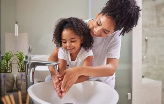 Imagem de uma mãe ajudando a filha a lavar as mãos em uma pia de banheiro para ilustrar matéria sobre a troca de titularidade Sabesp