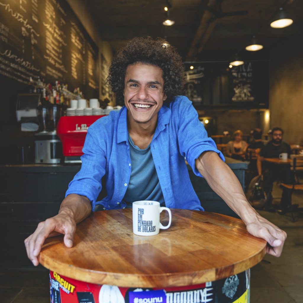 Foto do Tom Vieira que está em pé e de sorriso aberto na cafeteria Por Um Punhado de Dólares