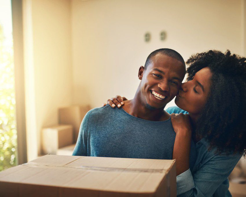 Foto que ilustra matéria sobre proposta de locação mostra um casal jovem que acaba de se mudar para uma casa nova. Na imagem, o homem sorridente ganha um beijo de sua parceira na bochecha.