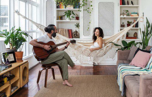 Foto mostra o casal Felippe e Carol na sala de seu apartamento no Paraíso, em São Paulo. Ele está sentado em um banco tocando violão, enquanto ela sorri sentada em uma rede de balanço