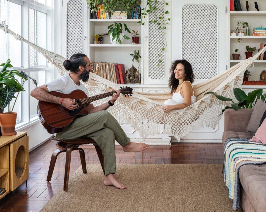 Foto mostra o casal Felippe e Carol na sala de seu apartamento no Paraíso, em São Paulo. Ele está sentado em um banco tocando violão, enquanto ela sorri sentada em uma rede de balanço