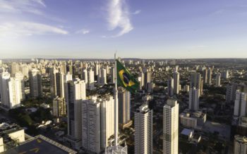 Foto que ilustra matéria sobre as melhores cidades para morar no Brasil mostra uma cidade repleta de prédios e uma bandeira do Brasil em primeiro plano, em um mastro no alto de um dos prédios.