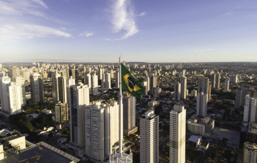 Foto que ilustra matéria sobre as melhores cidades para morar no Brasil mostra uma cidade repleta de prédios e uma bandeira do Brasil em primeiro plano, em um mastro no alto de um dos prédios.