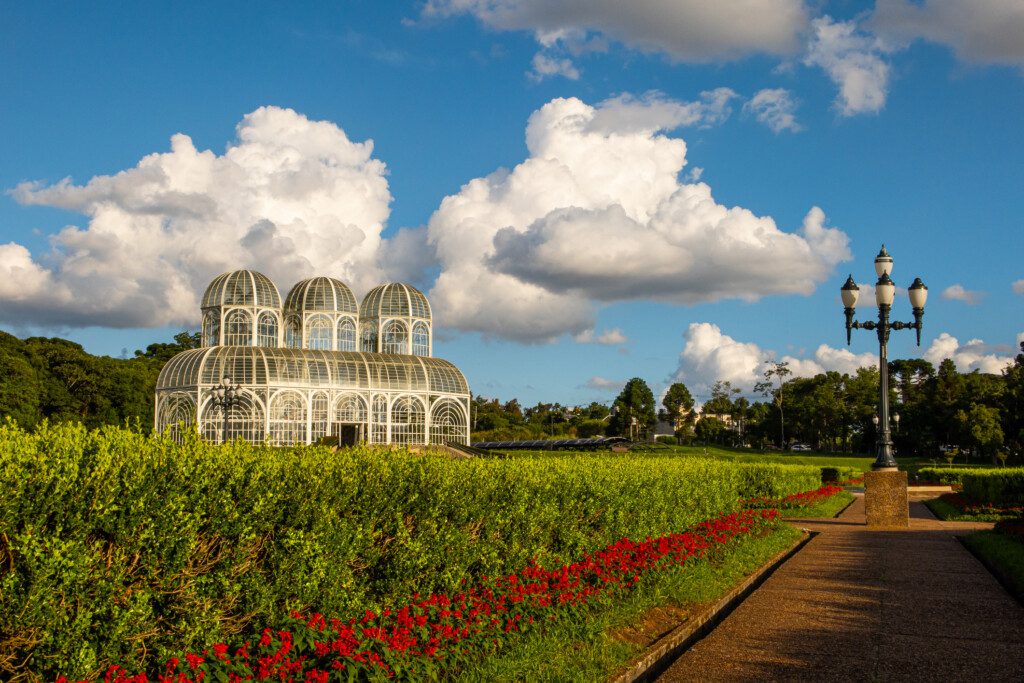 Imagem do Jardim Botânico, em Curitiba, um parque com jardins geométricos e uma estufa de vidro ao fundo