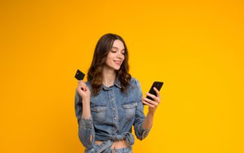 Foto que ilustra matéria que explica como pagar aluguel com cartão de crédito mostra uma mulher jovem olhando para o celular com um cartão de crédito na mão, com um fundo amarelo atrás dela.