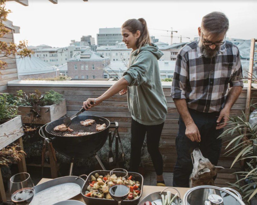A foto mostra uma área gourmet pequena. Há na imagem: um homem e uma mulher fazendo churrasco. Tem também uma churrasqueira, uma mesa com comidas e plantas decorativas.