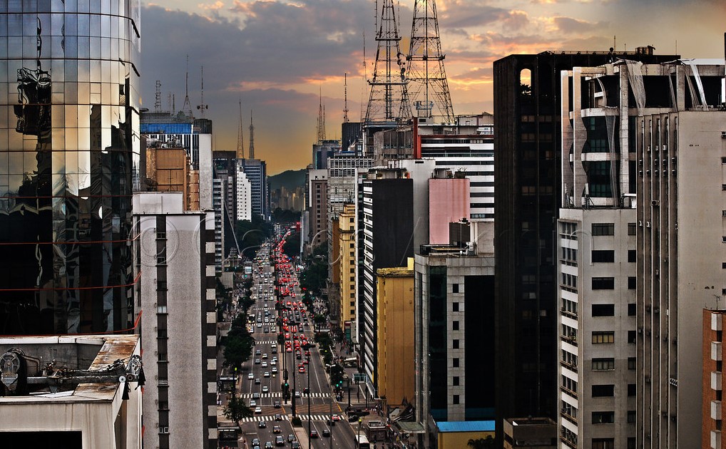 Avenida Paulista em uma imagem aérea no entardecer. Vemos os prédios que cobrem avenida e o trânsito de carros e ônibus