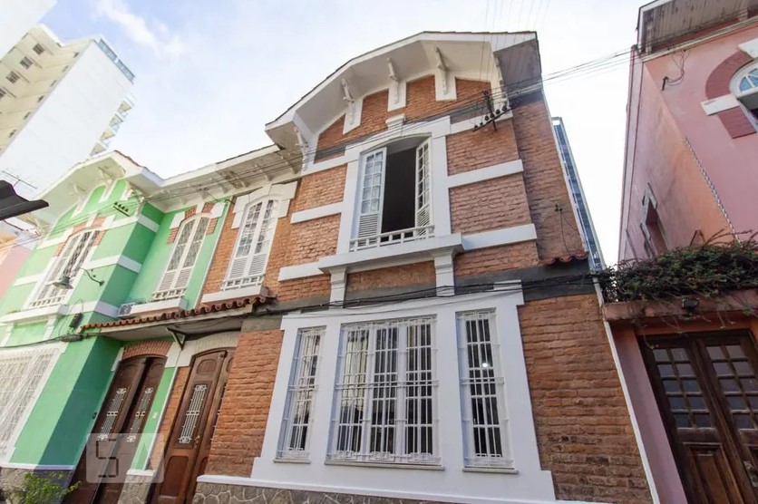 Casa com estilo arquitetônico colonial, com dois andares, uma janela ampla no primeiro andar. Material de tijolos marrons. Ao lado, há uma casa de tijolos verdes.