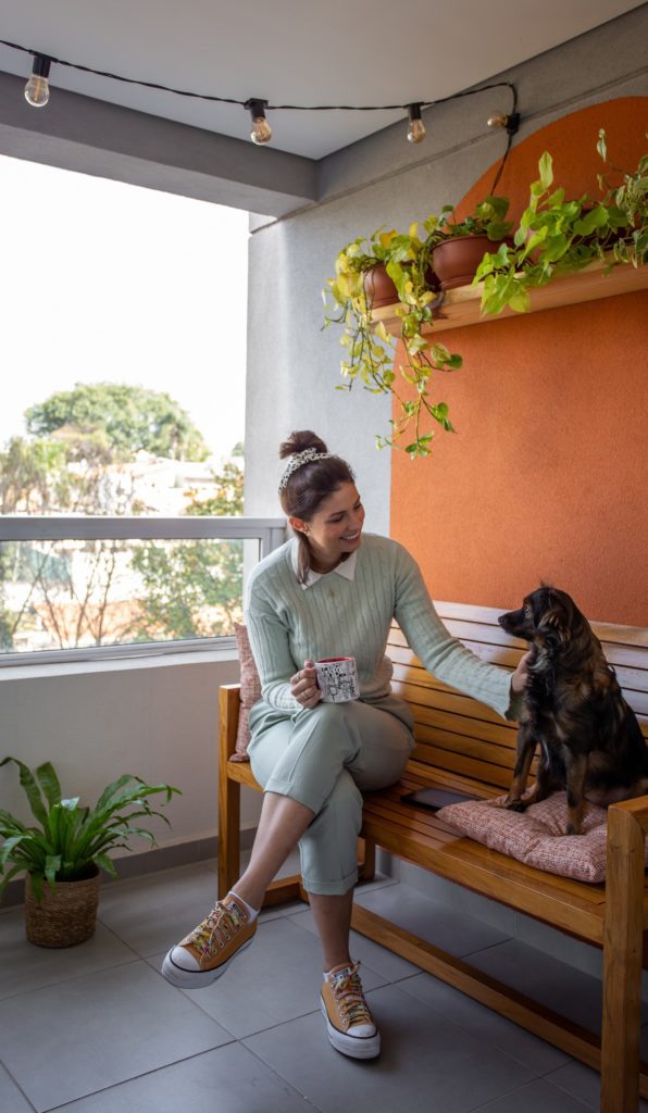 Raissa aparece segurando uma xícara e sentada em um banco na varanda com sua cadela Ana durante o dia.