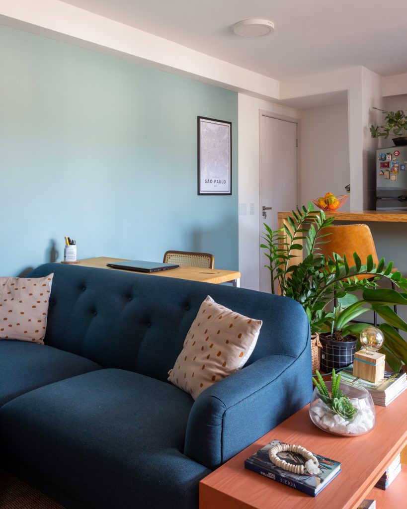 Foto mostra parte de uma sala, com um sofá azul em primeiro plano, uma mesinha com um laptop em cima e uma cadeira atrás e um detalhe da bancada de madeira da cozinha americana
