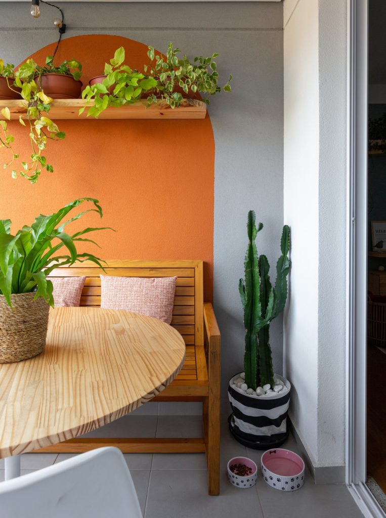 Foto que ilustra matéria sobre apartamento no Jardim Prudência mostra metade de uma mesa redonda com um vaso de plantas em cima, um banco de madeira ao fundo, com almofadas, um vaso com um cactus ao lado e uma parede laranja.