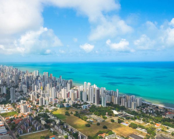 Foto que ilustra matéria sobre os melhores bairros de Recife mostra a cidade vista do alto, com muitos prédios no canto esquerdo da imagem e um mar azul cristalino mais à direita.