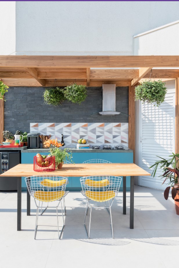 Área gourmet pequena com móveis e utensílios de cozinha, com uma área de churrasqueira em cores alegres, com plantas penduradas em suporte de madeira