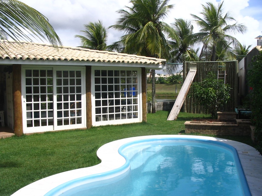 imagem de uma área gourmet externa com piscina rodeada por um gramado bem aparado. Aos fundos há uma cerca e palmeiras.