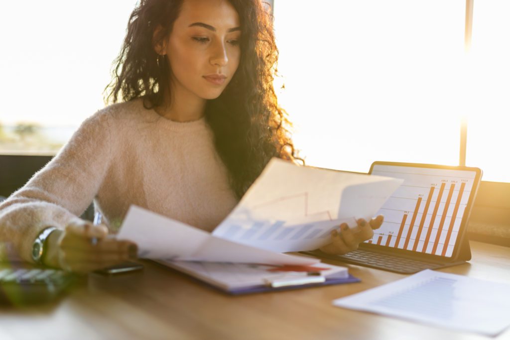 mulher analisando planilhas em dois papéis, com um notebook ao lado, ligado, mostrando gráficos, simulando o cálculo dos rendimentos imobiliários