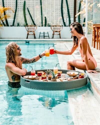 Um homem dentro da piscina brindando com uma mulher sentada à beira da água, ambos sorrindo. Entre eles há uma bandeja de frutas e coquetéis.