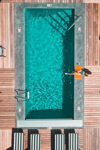 mulher de biquini verde à beira da piscina com o pé na água, sentada em uma toalha laranja