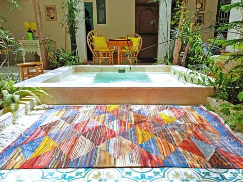 um tapete colorido à frente de uma piscina retangular. Aos fundos, duas cadeiras com almofadas amarelas, uma mesa e uma varanda.
