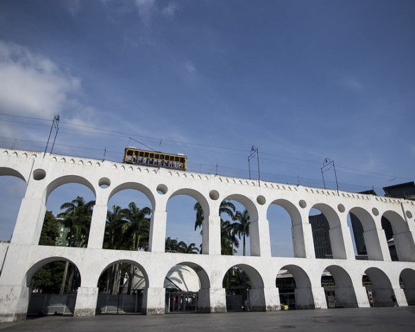 Foto que ilustra matéria sobre o Centro do Rio de Janeiro mostra a os Arcos da Lapa com o bondinho passando acima (Foto: Bruna Prado - MTUR)