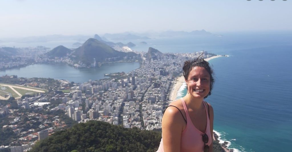 Foto que ilustra matéria sobre o Centro do Rio de Janeiro mostra a personagem da história, a relações públicas Lívia Abdalla, sorrindo, no alto de um morro, com uma vista do Rio de Janeiro ao fundo
