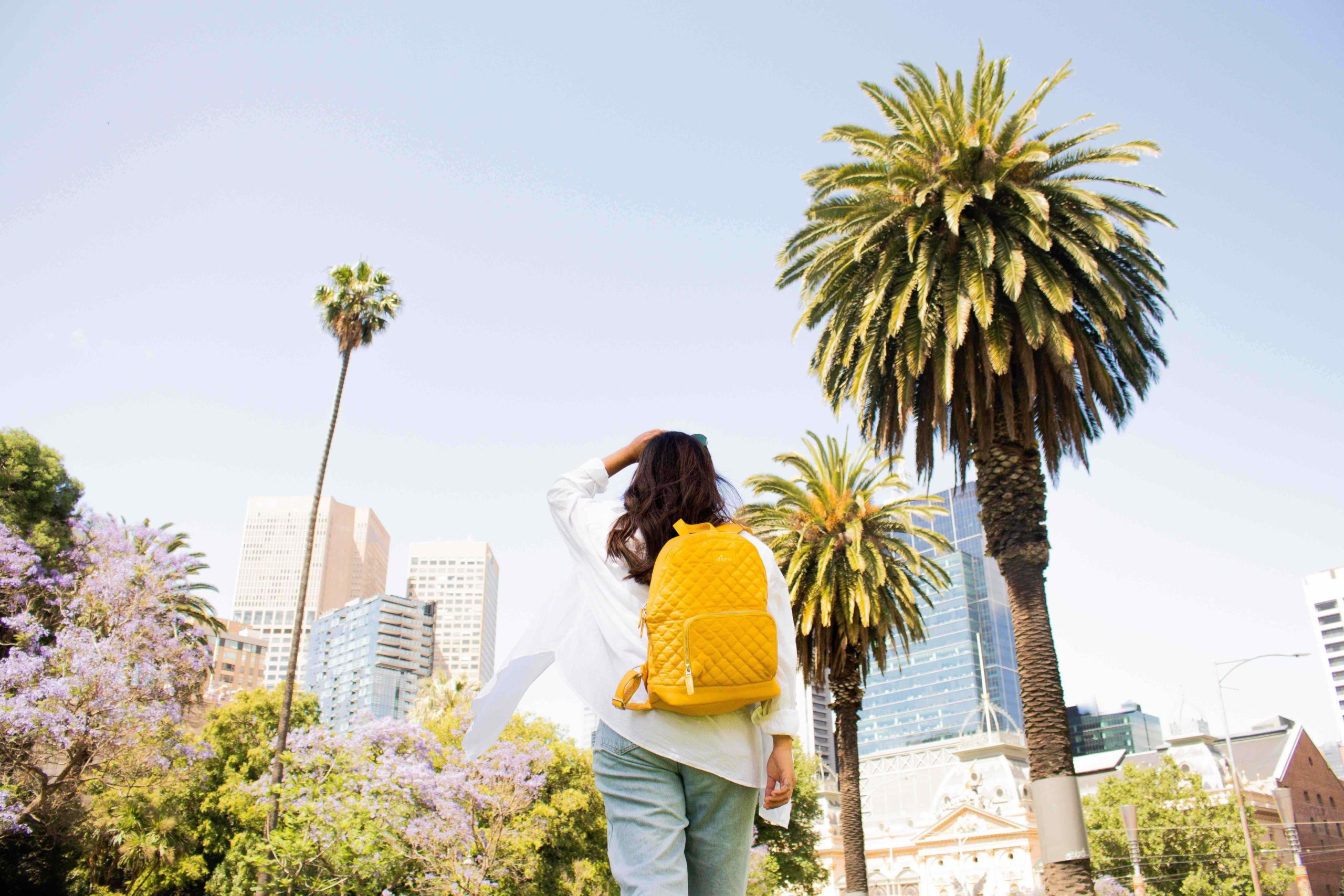 uma garota de mochila amarela de costas para a câmera, de frente para um conjunto de construções urbanas. Nas laterais da imagem há bastante folhagem, palmeiras, plantas e mais. 