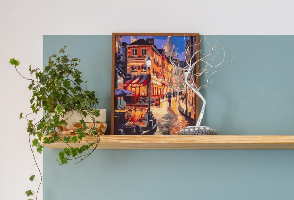 Foto que ilustra matéria sobre apartamento no Jardim Prudência mostra o detalhe de um quadro com uma pintura, posicionado em cima de uma prateleira com uma planta ao lado.