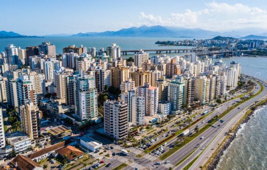 Foto que ilustra matéria sobre custo de vida em Florianópolis mostra uma visão aérea de vários prédios da cidade à beira-mar em um dia ensolarado, com uma ponte ao fundo.