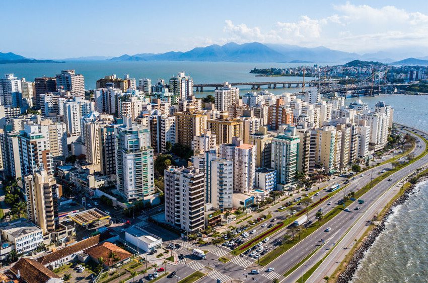 Foto que ilustra matéria sobre custo de vida em Florianópolis mostra uma visão aérea de vários prédios da cidade à beira-mar em um dia ensolarado, com uma ponte ao fundo.