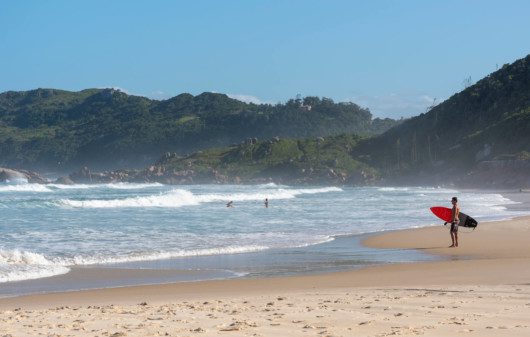 Foto que ilustra matéria sobre as praias de Florianópolis mostra um surfista de pé na areia, à direita da tela, com uma prancha de surfe debaixo do braço olhando para o mar na Praia da Galheta