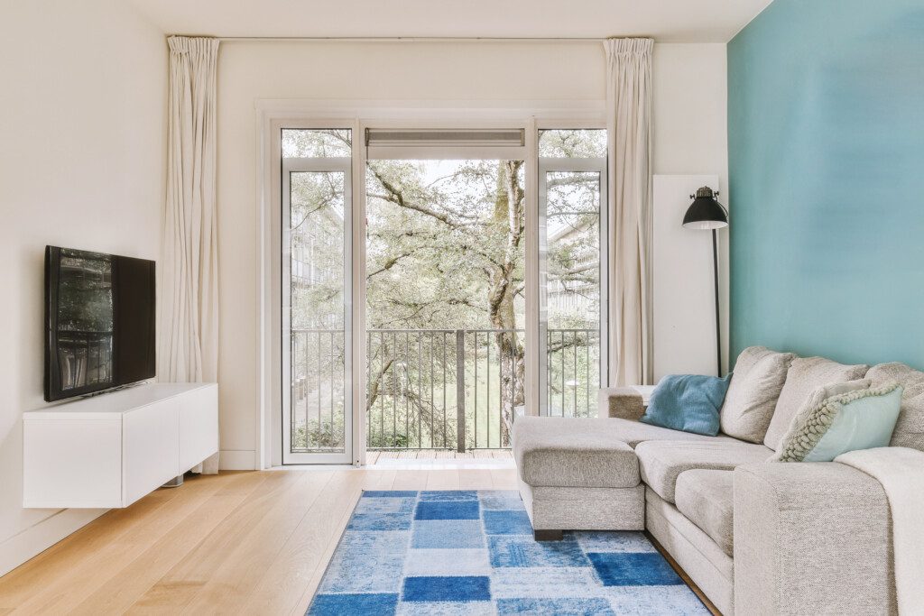 Sala de estar com grandes janelas e paredes brancas mobiliada com sofá confortável e puff em cima do tapete.