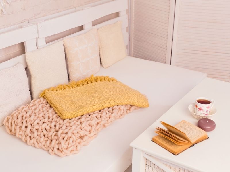 Sofá de palete branco com cobertores em tons pasteis aconchegantes e almofadas em tonalidades pasteis. Na frente, uma mesinha de centro com um café e um livro