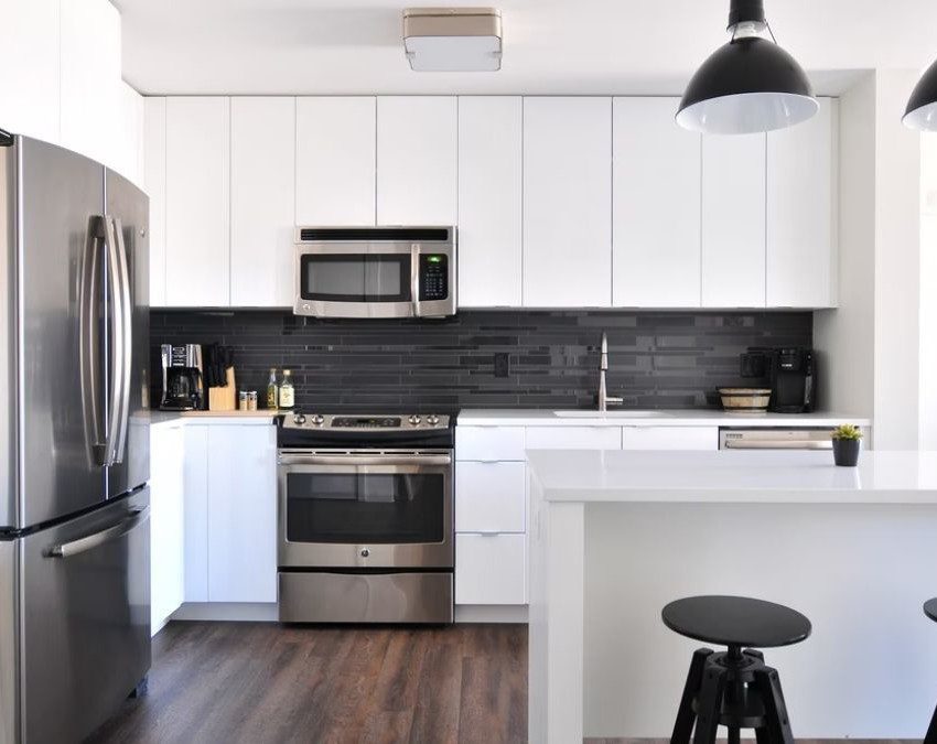 A foto mostra uma cozinha americana, com geladeira à esquerda, e fogão com pia e armários ao fundo. No lado direito, balcão com banquetas.