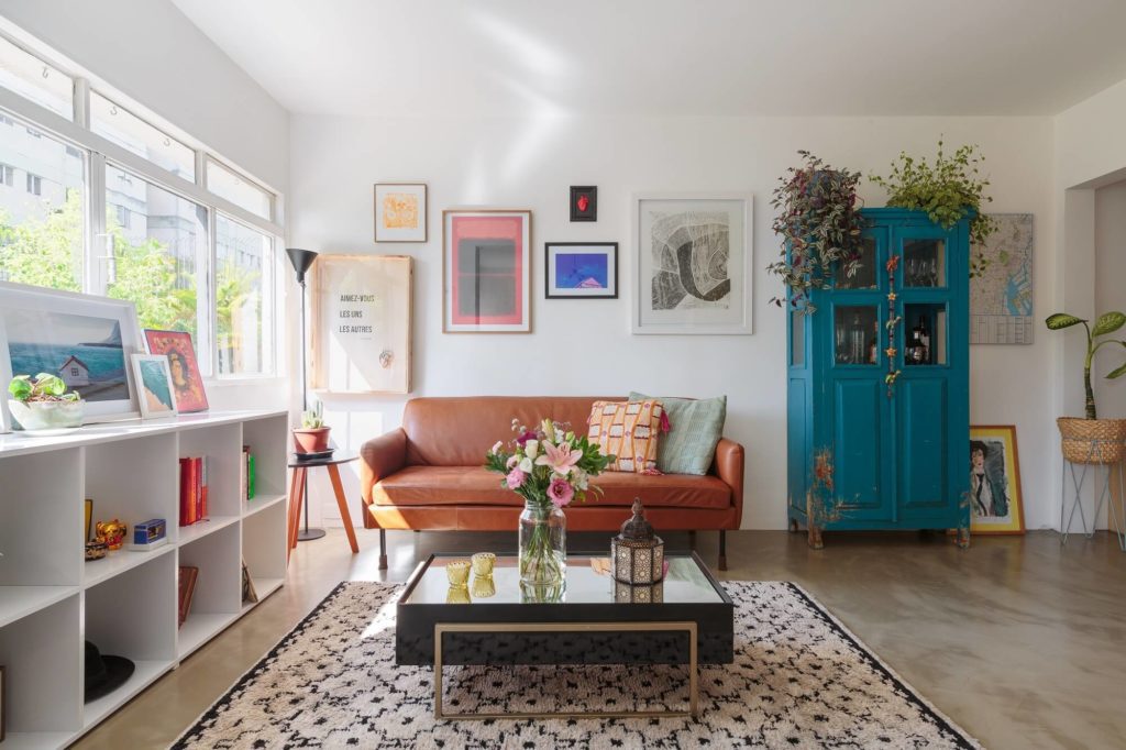 Foto que ilustra matéria sobre apartamento em Pinheiros mostra uma sala com um tapete e uma mesa de centro com tampo de vidro no meio. À direita, uma cristaleira azul com plantas em cima. À esquerda, uma grande janela e uma estante branca. Ao fundo, no centro, um sofá de couro marrom claro e quadros na parede.