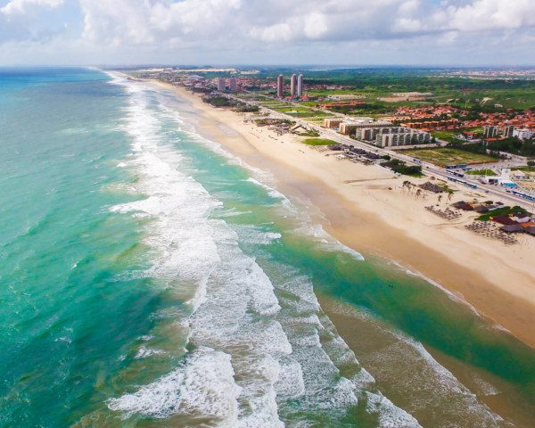 Foto que ilustra matéria sobre praias de Fortaleza mostra, de um ângulo do alto, boa parte da extensão da Praia do Futuro, com o mar aparecendo ao lado esquerdo da tela e a faixa de terra, com areia, algumas casas e vegetação, ao lado direito. (Foto: Jade Queiroz | MTUR)