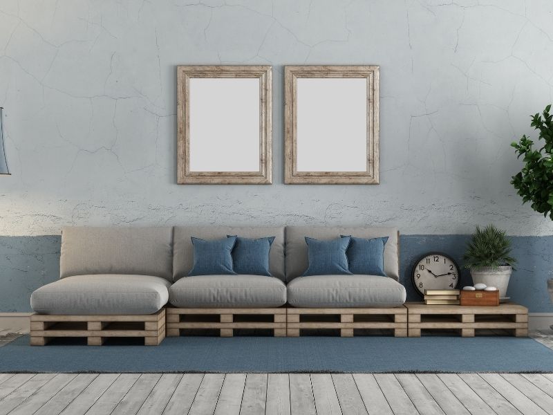Sofá de palete com três lugares, almofadas brancas e duas azuis, e dois espelhos na parede atrás dele. 