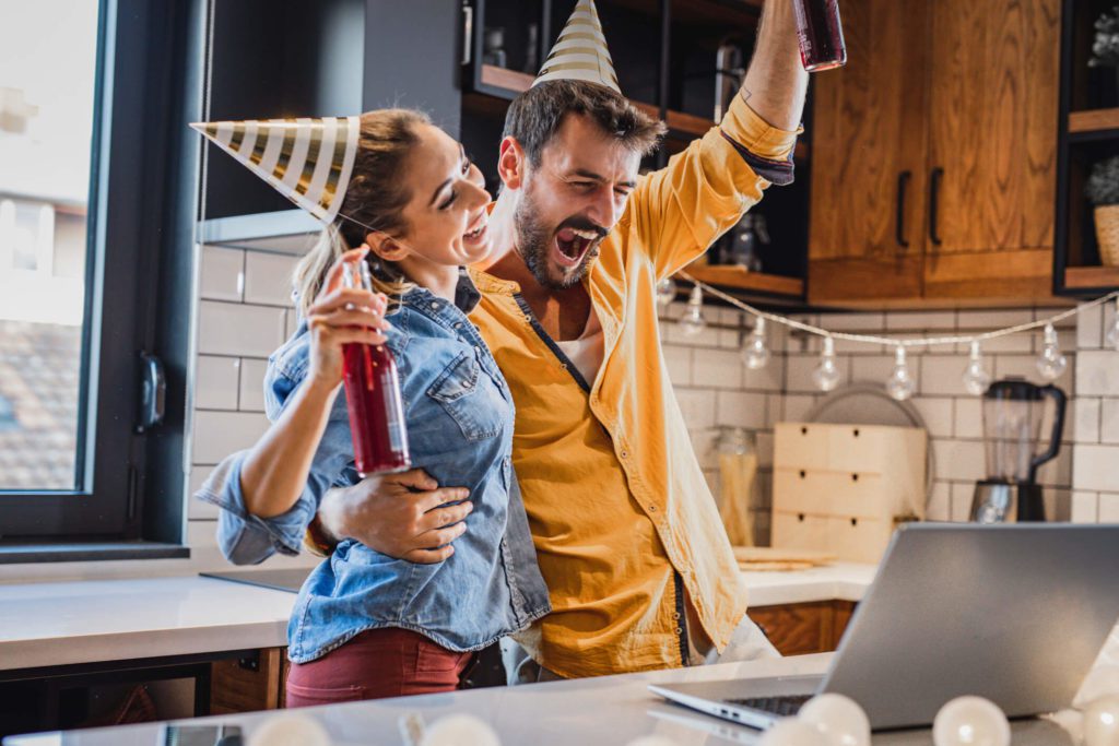 Foto que ilustra matéria sobre chá de casa nova mostra um casal em uma cozinha com um laptop em cima de uma bancada. Ambos de pé de frente para o computador, a mulher e o homem seguram garrafas de bebida, usam chapéus de festa e olham para a tela sorrindo.