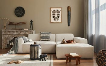 Foto que ilustra matéria sobre cores de parede para sala mostra uma sala com uma parede bege ao fundo, um sofá branco à frente e diversos objetos de decoração, tanto na parede quanto em uma estante entre a parede e o sofá.