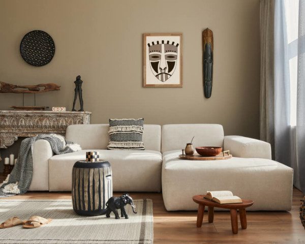 Foto que ilustra matéria sobre cores de parede para sala mostra uma sala com uma parede bege ao fundo, um sofá branco à frente e diversos objetos de decoração, tanto na parede quanto em uma estante entre a parede e o sofá.