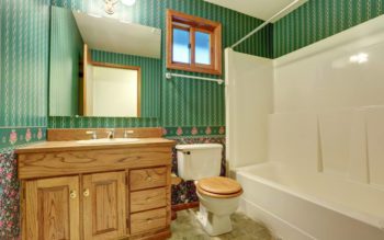 Foto que ilustra matéria sobre papel de parede para banheiro mostra um banheiro com um armário embutido de madeira sob a pia, um vaso sanitário com tampa de madeira clara e ao lado uma banheira. Ao fundo, a parede tem um papel de parede verde com listras, um espelho e uma pequena janela.