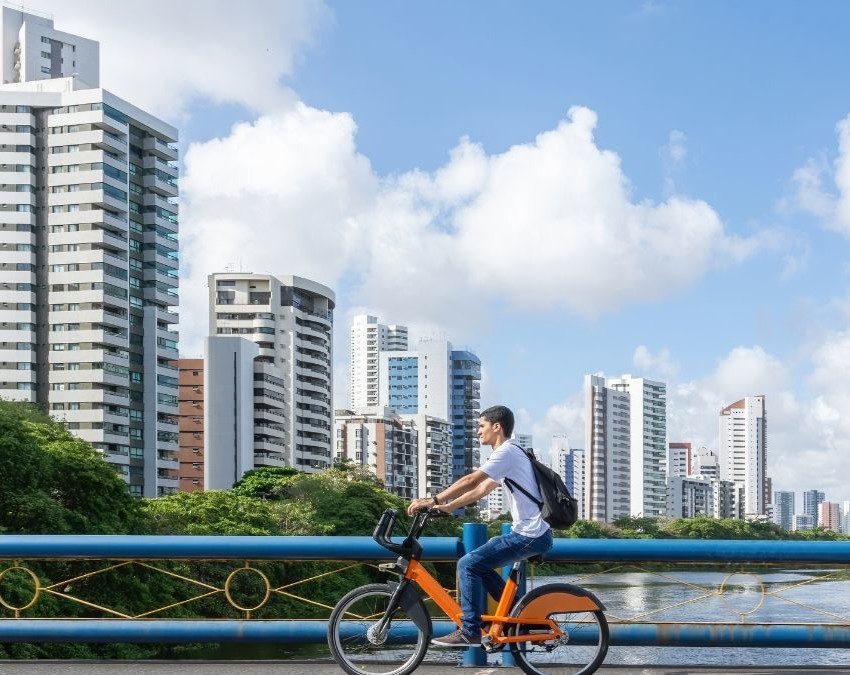homem andando de bicicleta às margens de um rio, com prédios na lateral esquerda da foto em Recife