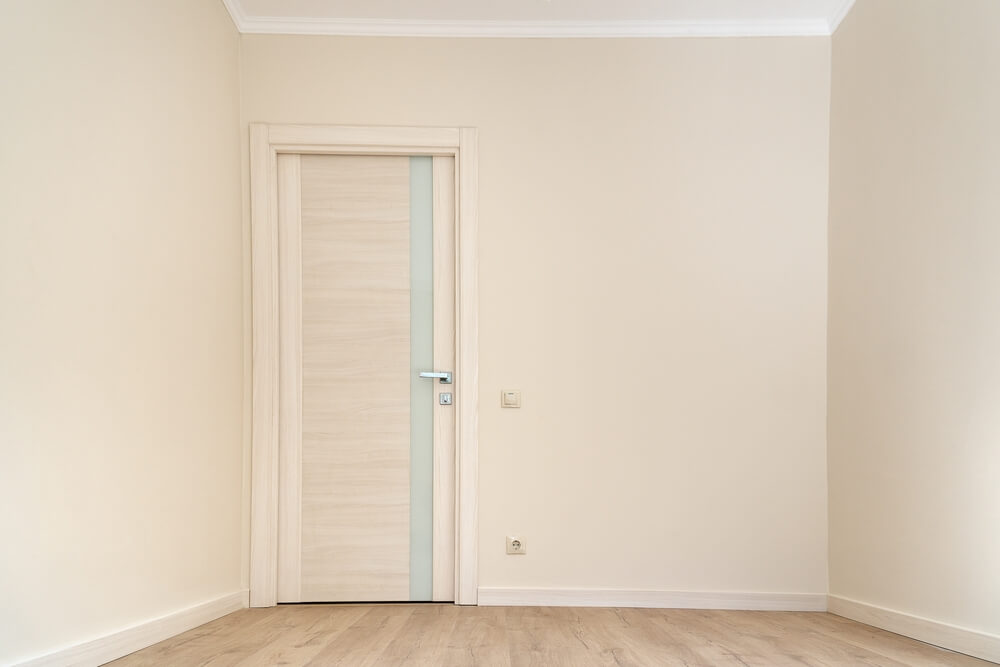 Foto que ilustra matéria sobre como colocar papel de parede mostra uma parede clara e vazia com uma porta mais à esquerda. A porta é coberta de um papel de parede em tom de madeira bem clara.